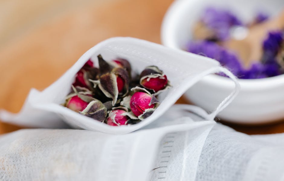 Close-up image showing a partially opened white paper bag filled with dried rosebuds, with some pink and dark red petals visible. In the background, there is a white bowl containing purple flower petals, placed on a wooden surface. The scene appears to be related to packing or preparing flora for removal or transport, consistent with a home relocation or packing process. The lighting is natural, highlighting the textures of the dried flowers and the paper bag. This image, associated with house removals services by Man With a Van Welling, illustrates the careful handling of delicate items during a furniture transport or packing and moving process within a property.