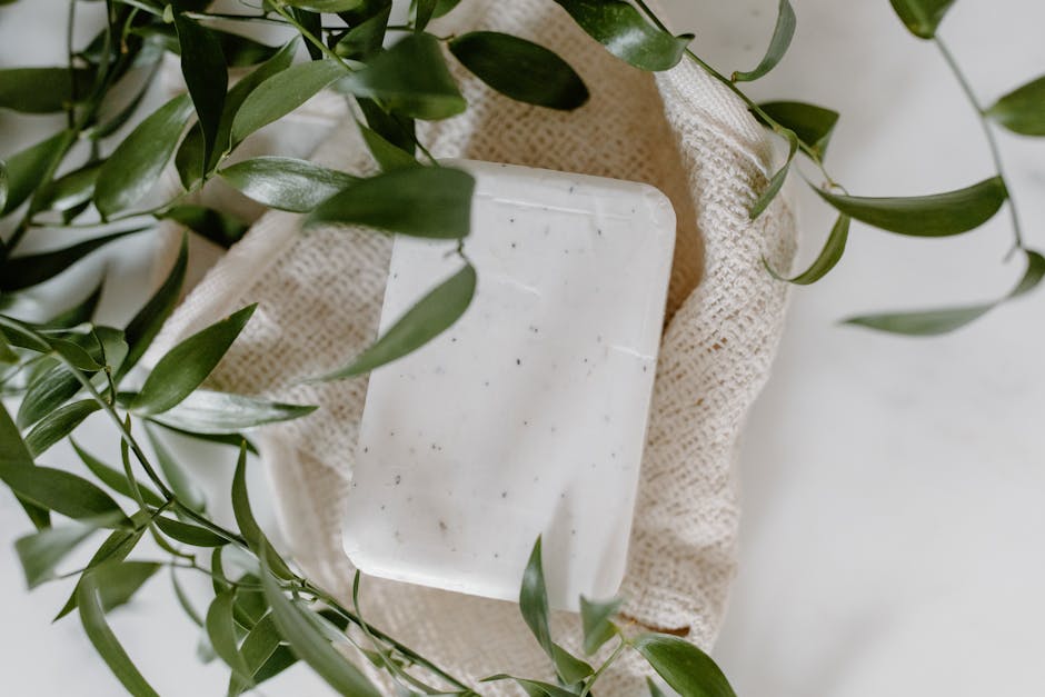A rectangular, white speckled soap dish made of ceramic or plastic rests inside a beige, textured fabric bag, which is partially open and placed on a white surface. Surrounding the bag and soap dish are green leafy plant stems, with some leaves draping over the objects. The scene is well-lit with natural light, creating soft shadows. This arrangement suggests a focus on household items and packing materials commonly involved in home relocation or moving services, as offered by Man With a Van Welling, emphasizing organized packing and the careful handling of household goods during a house move.