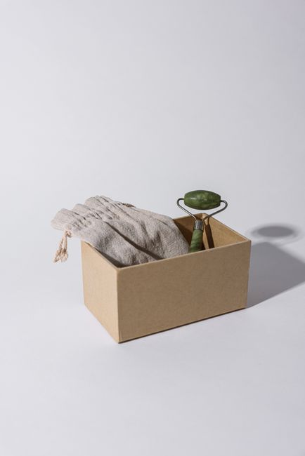 Close-up image showing a partially opened white paper bag filled with dried rosebuds, with some pink and dark red petals visible. In the background, there is a white bowl containing purple flower petals, placed on a wooden surface. The scene appears to be related to packing or preparing flora for removal or transport, consistent with a home relocation or packing process. The lighting is natural, highlighting the textures of the dried flowers and the paper bag. This image, associated with house removals services by Man With a Van Welling, illustrates the careful handling of delicate items during a furniture transport or packing and moving process within a property.