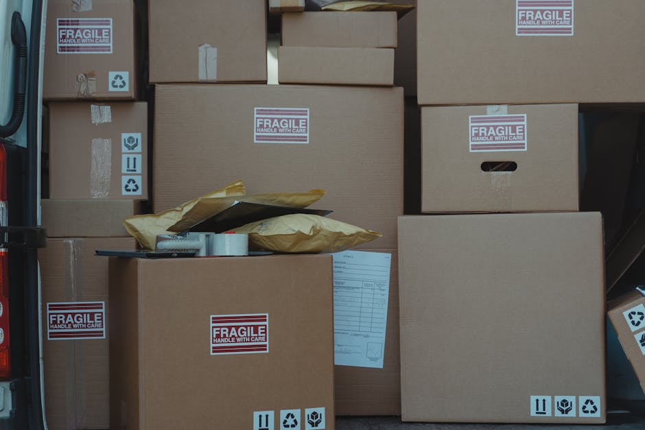 A side view of a vehicle trunk loaded with multiple brown cardboard boxes, some wrapped in light brown paper with yellow packing paper, and marked with sticky labels reading 'FRAGILE HANDLE WITH CARE.