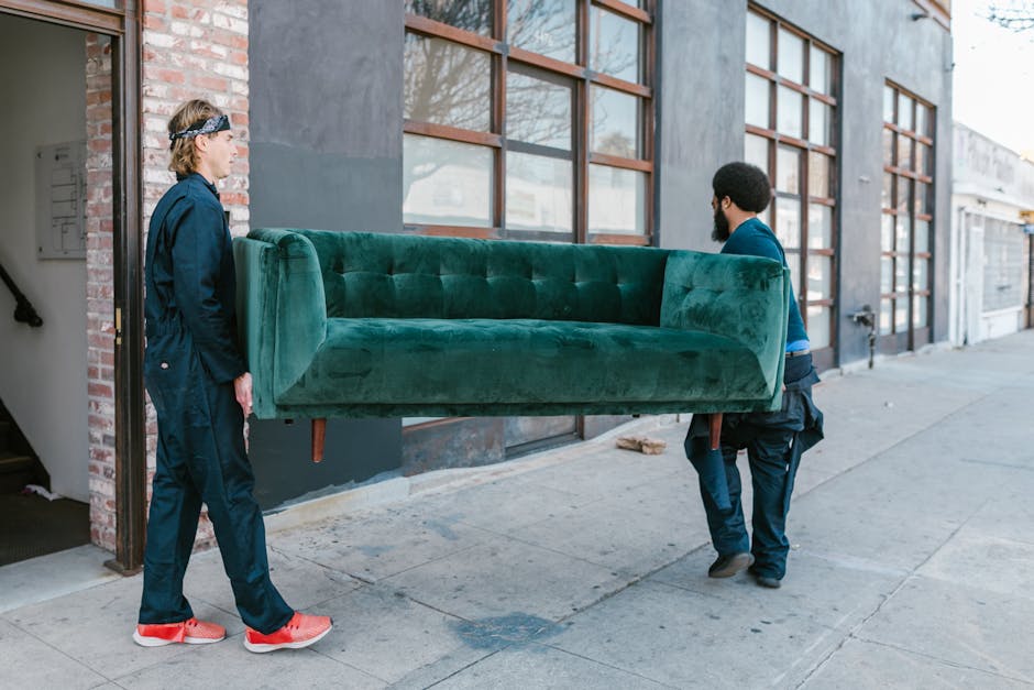 Two men are engaged in a home relocation process outside a commercial building with large windows and a brick wall on the left. One man, wearing a black jacket, navy trousers, and red shoes, is standing on the sidewalk, observing as the other man, dressed in a blue shirt and dark trousers, carefully carries a large, green velvet sofa with wooden legs. The sofa is positioned horizontally between them, indicating a furniture transport activity related to packing and moving. The scene suggests the loading process of furniture for a house removal, with the men handling the sofa with care, possibly preparing it for transport in a van parked nearby. The environment is well-lit, with natural daylight highlighting the outdoor setting, which is typical for moving services offered by companies like Man With a Van Welling.