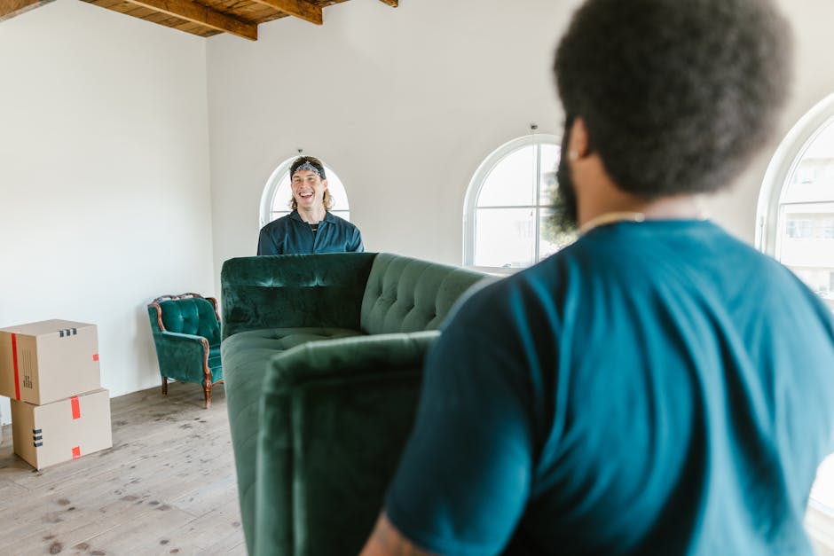 Inside a bright, white-walled room with two large arched windows allowing natural daylight, a professional moving team from Man With a Van Welling is conducting a home relocation. A man with dark, curly hair and a blue shirt is seen from behind, holding a rolled-up green velvet sofa that is part of the furniture being moved. In front of him, a woman with light skin, long hair, and wearing a safety headband and dark top, is smiling as she guides the furniture through the space. To the left, two cardboard boxes with red and black tape are stacked on the wooden floor, indicating packing processes, while a small vintage-style green armchair is positioned near the window, awaiting transport. The scene captures the careful handling of furniture and the organized logistics involved in packing and moving a house, with the environment set for a professional relocation service by Man With a Van Welling, ensuring smooth furniture transport and effective moving logistics.