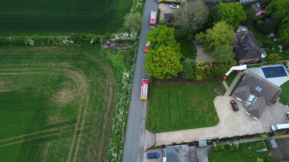 An aerial view of a residential property showing a driveway with a red and white removal van from Man With a Van Welling parked at the entrance. The house has a modern design with a grey roof, white exterior walls, and a blue swimming pool in the backyard. Surrounding the house are landscaped gardens, including neatly trimmed grass, bushes, and mature trees with green foliage. Several cardboard boxes and packing materials are visible on the driveway indicating packing activities. The van is positioned to load or unload furniture and boxes as part of a home relocation or removal service. Adjacent to the driveway, there is a paved pathway leading to the house entrance, and other parked cars are seen along the street. The environment is well-lit with natural daylight, clearly showing the process of loading household items into the van as part of an ongoing moving process, typical for emergency same-day removals in Welling.