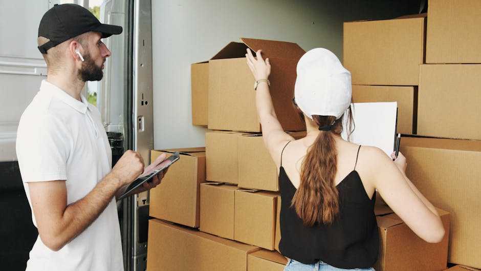 A man and woman are engaged in a house removal or furniture transport process next to an open moving van. The man, wearing a black cap, white polo shirt, and white earbuds, holds a clipboard and pen, inspecting the packing. The woman, dressed in a white cap, black sleeveless top, and jeans, is seen from behind, reaching over a stack of large cardboard boxes with white packing paper visible at the edges. The boxes are arranged on the ground and inside the van, which has its rear doors open, revealing more packed items. The setting appears to be on a driveway or pavement adjacent to a property, with natural daylight illuminating the scene. The environment suggests a home relocation involving packing, loading, and transportation of household goods, with the presence of trolleys or straps not visible but implied in the process, supporting the services of Man With a Van Welling in quick, professional removals. The focus is on the logistical steps involved in packing and readying furniture and boxes for moving.