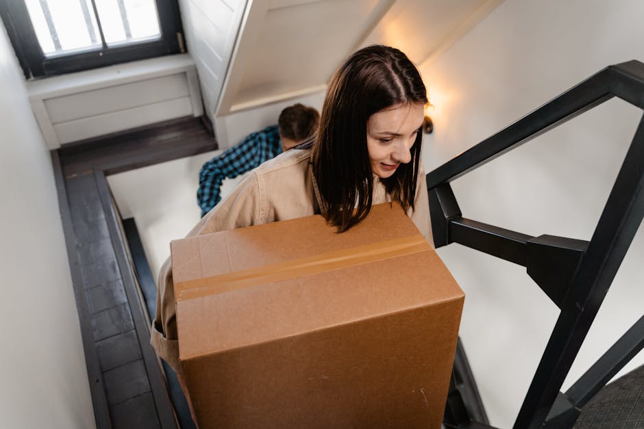 A woman with shoulder-length dark hair carrying a large cardboard box, wrapped with packing tape, up a narrow staircase inside a residential property. Behind her, a man in a blue checkered shirt is visible, also climbing the stairs with additional packed boxes. The staircase has dark wooden steps, white walls, and a black handrail on one side. Natural light is coming through a window next to the stairs, illuminating the scene. The interior shows signs of packing and home relocation, with several boxes likely prepared for moving. Man With a Van Welling, a professional removals company, is involved in handling the furniture transport and packing process to facilitate moving from a Welling flat with tight stairs, ensuring the safe and efficient handling of household items during the home relocation.