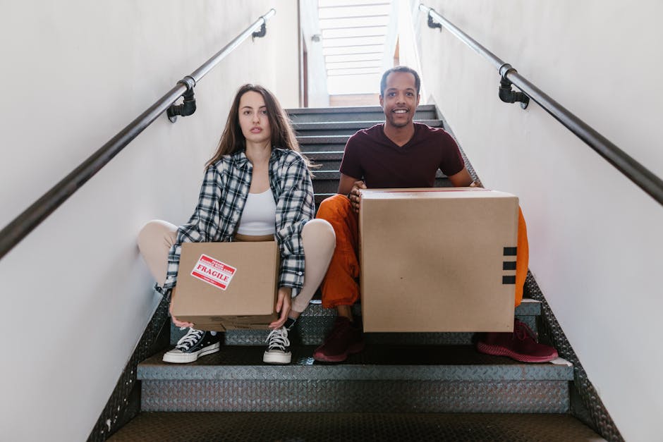 Two movers from Man With a Van Welling are positioned on a staircase inside a property, preparing for a home relocation. The female mover, seated on the stairs, holds a small cardboard box labeled 'fragile' with red and white stickers, dressed in casual clothing and sneakers. The male mover, beside her, is standing and holding a larger cardboard box, wearing a maroon T-shirt and orange trousers. The staircase features metal railings on both sides, with natural light coming from a window or doorway at the top, illuminating the scene. The background shows white walls and a staircase with grey carpeted steps, indicative of an internal moving process in a flat or apartment setting. This image visually depicts the logistics of packing and loading furniture and boxes during a professional move, emphasizing careful handling and the importance of efficient transport within a property.