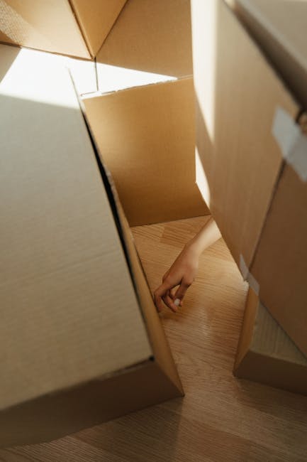 A woman with shoulder-length dark hair carrying a large cardboard box, wrapped with packing tape, up a narrow staircase inside a residential property. Behind her, a man in a blue checkered shirt is visible, also climbing the stairs with additional packed boxes. The staircase has dark wooden steps, white walls, and a black handrail on one side. Natural light is coming through a window next to the stairs, illuminating the scene. The interior shows signs of packing and home relocation, with several boxes likely prepared for moving. Man With a Van Welling, a professional removals company, is involved in handling the furniture transport and packing process to facilitate moving from a Welling flat with tight stairs, ensuring the safe and efficient handling of household items during the home relocation.