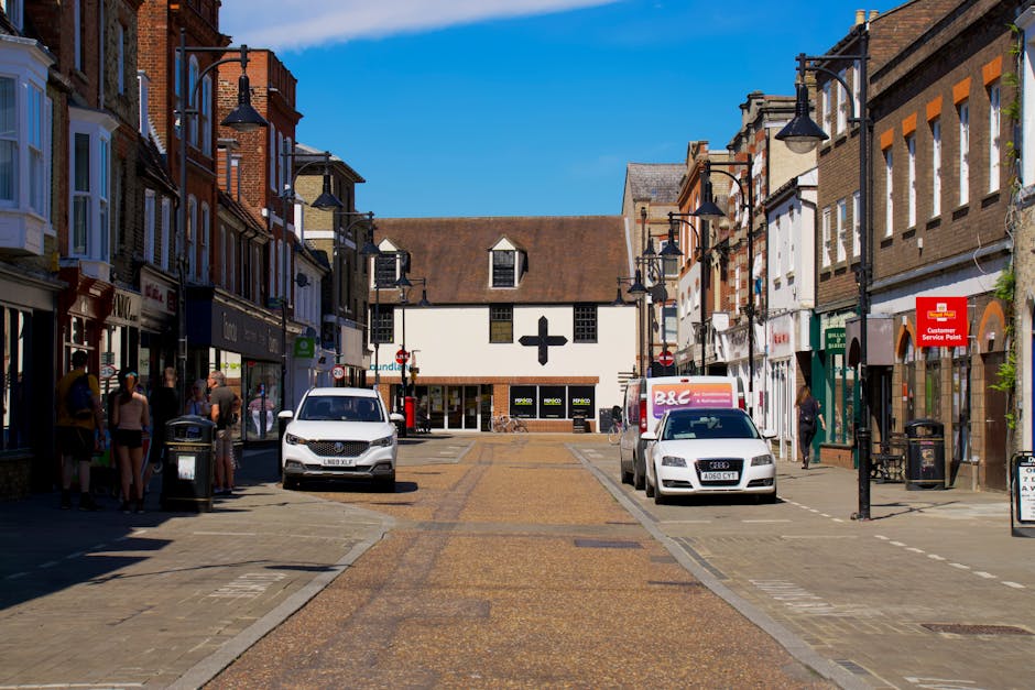 A street scene on Welling High Street showing a typical high street with brick and painted shopfronts on both sides, under a bright blue sky. The image captures a home relocation process with two white vans parked along the pavement, one of which is branded with packaging and furniture transport materials for moving services by Man With a Van Welling. The vans are positioned near the curb, with parts of furniture, cardboard boxes, and packing materials like plastic wraps visible on the pavement and inside the vehicles, indicating preparations for a furniture transport or full house removal. Several pedestrians are walking or standing nearby, and the street features traditional lampposts, a pharmacy sign, and a customer service point sign attached to the buildings. This scene reflects the practical logistics involved in packing and moving, with the loading process beginning beside commercial premises, supporting effective house removals and professional moving services in Welling.