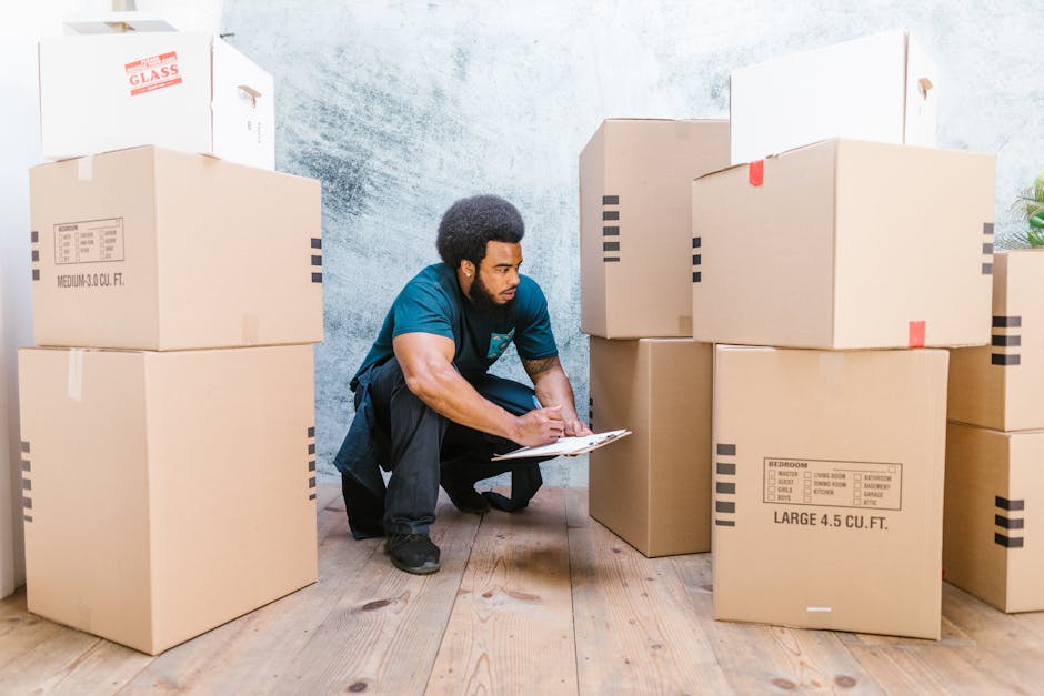 A man with curly black hair, wearing a blue t-shirt and black trousers, is kneeling on a wooden floor inside a property, surrounded by large cardboard moving boxes labeled with room contents such as b