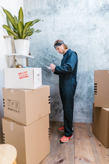 A man with curly black hair, wearing a blue t-shirt and black trousers, is kneeling on a wooden floor inside a property, surrounded by large cardboard moving boxes labeled with room contents such as b