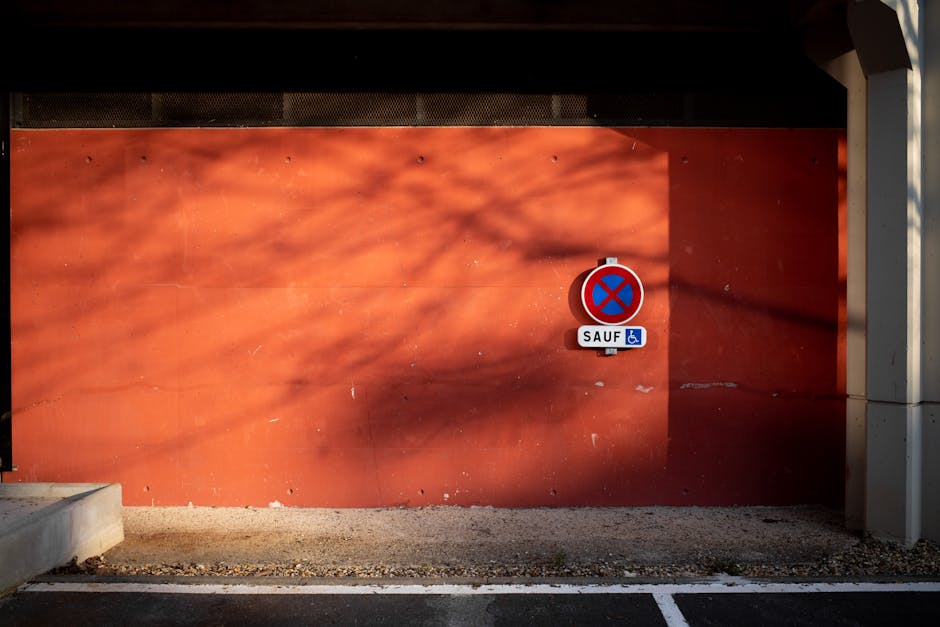 The image shows a solid orange wall adjacent to a residential property, with a parking restriction sign attached to the center of the wall indicating no waiting or parking, including exceptions for disabled access symbols. The sign is mounted on a small metal pole, and the ground beneath the wall consists of gravel and asphalt pavement. To the left, part of a concrete step leading to an entryway is visible, while on the right, a white structural column supporting an overhanging roof casts a shadow on the wall, creating a contrast with sunlight. The scene is illuminated by natural daylight, highlighting the textures of the wall, pavement, and nearby structural elements. This setting appears to be part of the exterior environment where home relocation or furniture transport might be planned, and the sign indicates parking restrictions relevant to moving logistics. The image reflects an outdoor space typical for loading or unloading during house removals performed by [COMPANY_NAME], such as Man With a Van Welling.