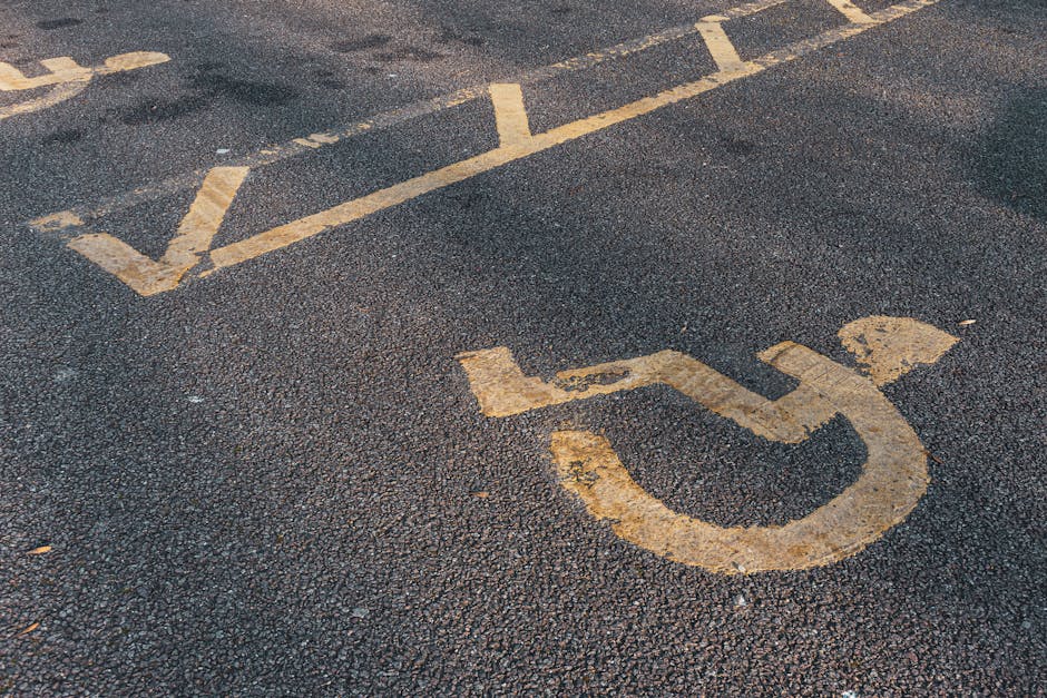 A close-up view of a designated accessible parking space on an asphalt surface, featuring a yellow painted wheelchair symbol and dashed lines indicating its boundary. The surface shows signs of wear with some fading and small cracks. In the background, there are additional parking lines, suggesting the space is part of a larger parking lot. The image provides context for parking, access, and permits related to household removals in Welling, as managed by Man With a Van Welling, supporting efficient home relocation, furniture transport, and packing and moving processes.