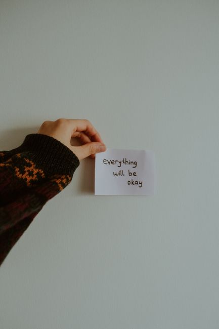 A person's hand, wearing a dark knitted sleeve with orange and brown patterns, holds a small white piece of paper with the handwritten message 'everything will be okay' against a plain, light-colored wall. The image is captured indoors, featuring no furniture or other objects, emphasizing the message's calming intent. This scene could be associated with emotional support during a home relocation or moving process, reflecting reassurance for homeowners. The natural lighting creates a soft, neutral atmosphere, and the image is relevant to moving or packing services provided by companies like Man With a Van Welling, supporting the theme of smooth and supportive home removals.
