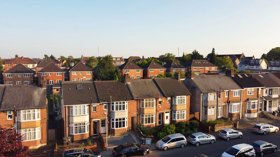 An aerial view of a residential street during daylight hours, showing a row of terraced houses with pitched roofs, some with skylight windows. A large green tree is centrally located, partially obscuring the view of some houses. Parked cars line the narrow street, which is paved with asphalt, and the pavement features small retaining walls and steps leading to house entrances. The scene takes place outside a typical UK home, with outdoor areas and driveways visible, illustrating surroundings where a professional home removal or furniture transport service like Man With a Van Welling might operate during the loading process of a house relocation. The natural lighting emphasizes the details of the houses, vehicles, and greenery, reinforcing the context of furniture transport and packing during a residential move.
