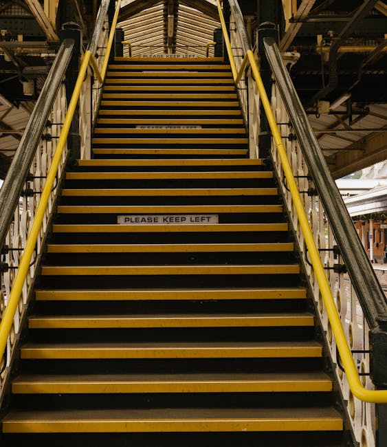 A set of industrial metal stairs with black and yellow striped treads leading upward inside a warehouse or loading area. The stairs feature yellow handrails on both sides and are positioned in front of a corrugated metal ceiling. Several signs on the steps display the message 'PLEASE KEEP LEFT,' indicating directional guidance for movement. Surrounding the staircase, there are parts of a vehicle, likely a van or truck used for home relocation, with visible wheels and loading equipment, such as straps and blankets, suggesting an active furniture transport or packing and moving process. The environment is well-lit with natural light possibly coming from openings or windows in the background. This scene reflects a professional removals setting, where careful handling and transportation of household items are underway, supported by the moving company Man With a Van Welling.