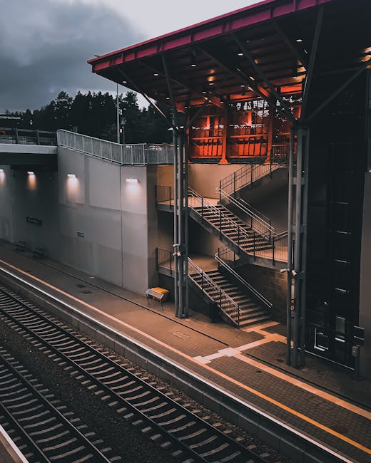 A nighttime view of an exterior stairway at Welling railway station, with metal railings and steps leading up to an overhead walkway enclosed in glass. The station platform, made of concrete with tactile paving and yellow safety lines, runs alongside railway tracks that extend into the distance. The station building features a red roof supported by metal beams, with a corner of the building visible in the image. External lighting illuminates the platform and stairs, while the dark sky indicates early evening or overcast weather. The scene reflects the typical setting of a suburban station used by local residents for home relocation or commuter travel, with the station's access structure and surrounding environment subtly highlighting logistical considerations related to moving and transportation services such as those offered by Man With a Van Welling.