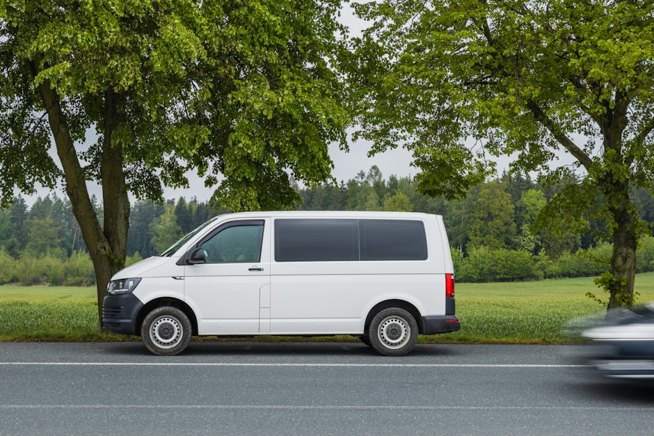 A white commercial van parked on the side of a paved road in front of a grassy field with lush green trees overhead. The van is positioned parallel to the roadside, with the driver’s side facing the viewer. It features a sliding door on the side, which is closed, and rear doors that are also shut. The van’s windows are tinted, and the vehicle appears clean and in good condition. Behind the van, a natural landscape with open fields and dense woodland extends into the distance. The scene is captured in daylight with bright, even lighting, and a slight motion blur of a passing vehicle on the right side suggests movement relevant to home relocation or moving logistics. The image visually supports transportation and loading processes involved in house removals, as carried out by companies like [COMPANY_NAME], and illustrates the type of vehicle suitable for furniture transport and packing and moving tasks related to [PAGE_TITLE].
